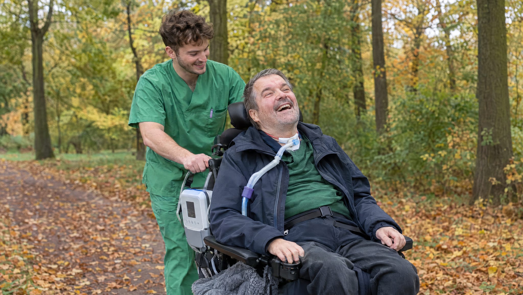 Begleiter schiebt einen lachenden Mann mit Beatmungsgerät im Rollstuhl durch den herbstlichen Park, Symbolbild, KI-generiert